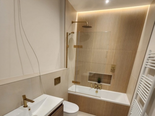 A modern bathroom renovation in Dublin featuring warm beige large-format tiles, a ribbed texture feature wall behind the bath, gold fixtures, and ambient LED lighting.