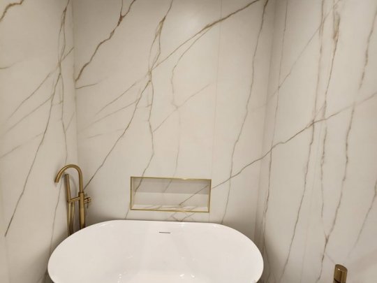 A luxury modern bathroom in Dublin featuring floor-to-ceiling white marble-effect porcelain tiles with gold veining, a freestanding bathtub, and gold brassware.