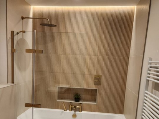 Modern bathroom with textured beige wall tiles, warm LED lighting, brass fixtures, glass shower screen, and white bathtub.