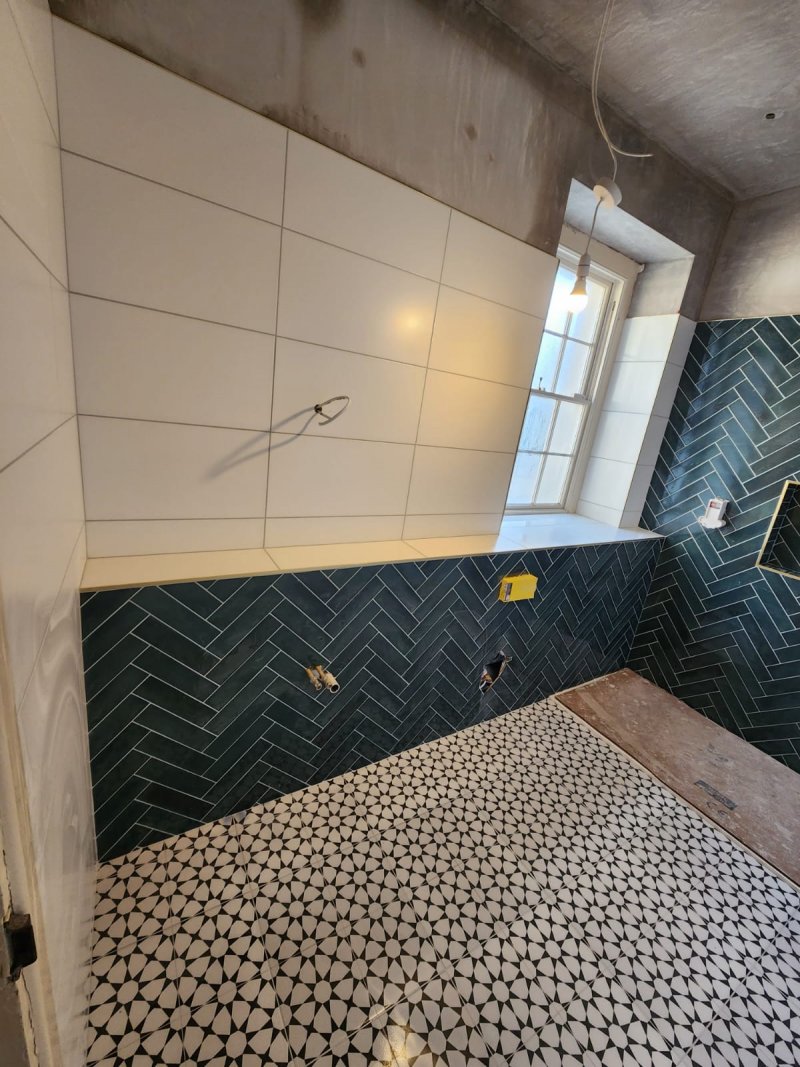 A bathroom during renovation, featuring striking black and white geometric floor tiles and a dark green herringbone-patterned feature wall with a built-in shower niche.