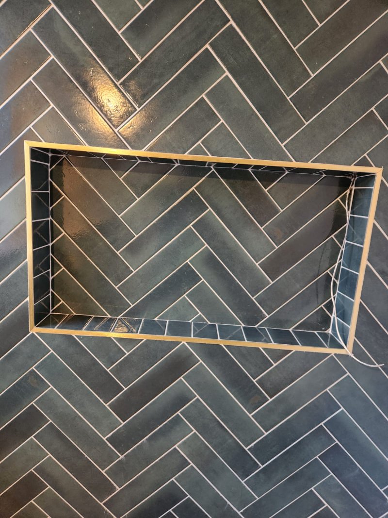 A close-up of a rectangular, brass-trimmed shower niche seamlessly integrated into a wall of dark green, herringbone-patterned tiles.