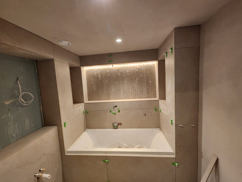 Modern bathroom tiling in progress by RCTiling Dublin, showing large beige tiles around a bathtub with an illuminated recessed niche and tile leveling system.