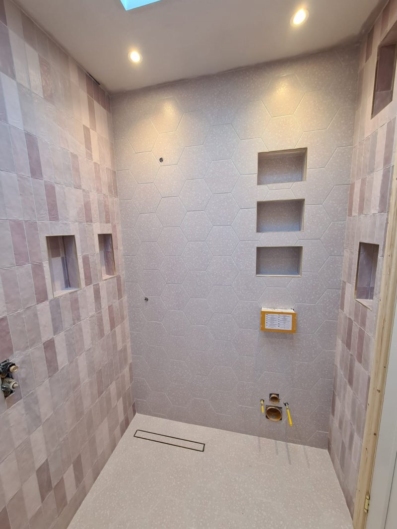 A walk-in shower under construction featuring a main wall with large, pale hexagonal tiles and three built-in niches, contrasted by a side wall with dusty rose vertical rectangular tiles.