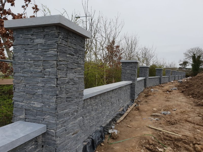 Long residential boundary wall clad in dark stacked stone with stone pillars and capping.