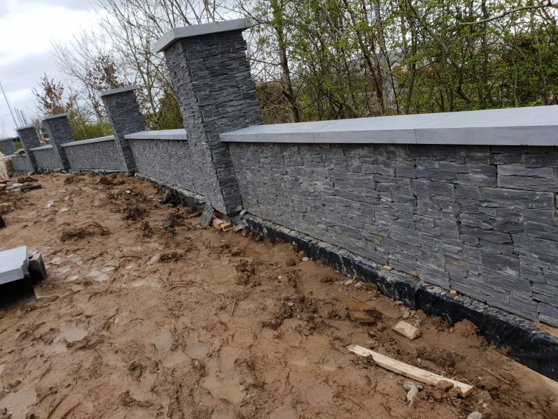 A long, residential boundary wall with dark stacked stone cladding, stone pillars, and a finished capping.